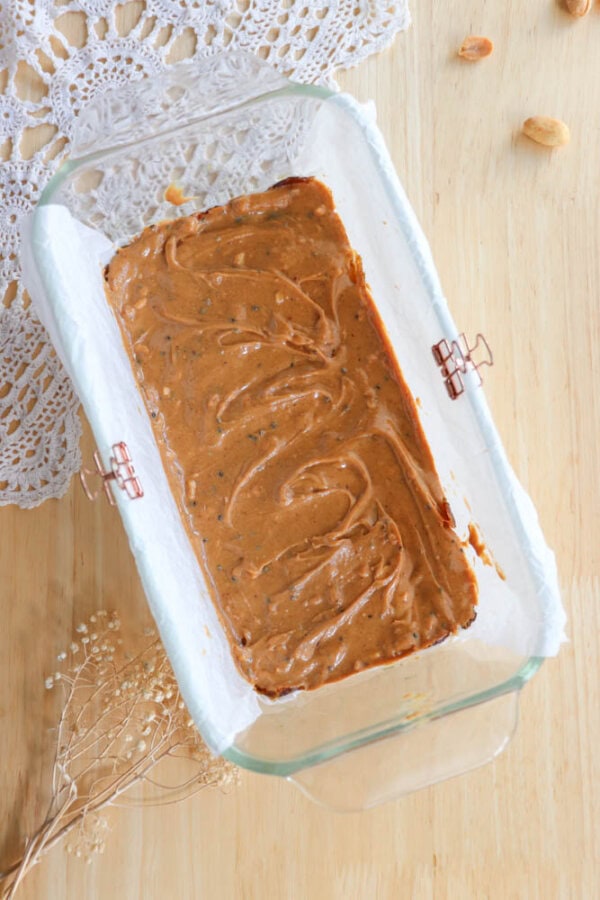 A glass baking dish lined with parchment paper with chocolate brownie batter spread evenly inside. The dish is placed on a wooden surface with a lace doily and some small flowers nearby.