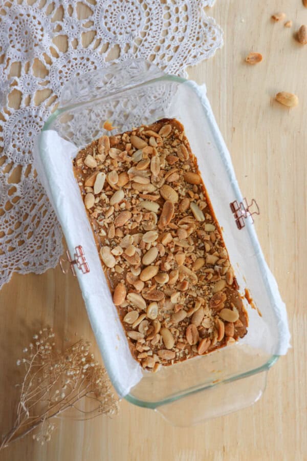 A glass baking dish lined with parchment paper contains a nut-topped dessert resting on a wooden table. Next to it is a lace doily and a small sprig of dried flowers. Some nuts are scattered around.