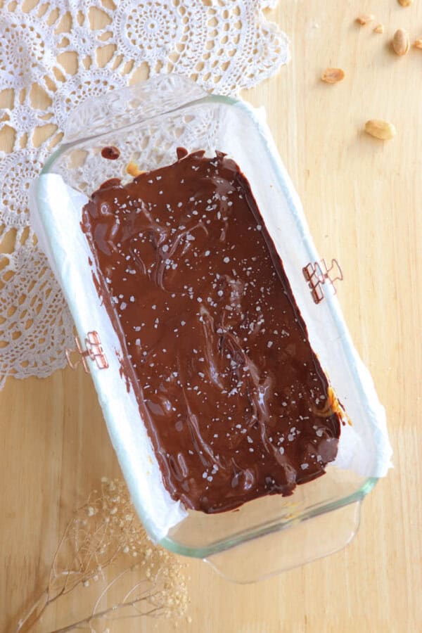 A glass dish lined with parchment paper holds homemade chocolate bark sprinkled with sea salt. The dish is placed on a wooden surface with a decorative lace cloth and a few scattered nuts nearby.