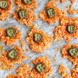 Close-up of baked cheese crisps with a slice of pickled green cucumber on each, resting on a parchment-lined baking sheet. The crisps are golden-brown and scattered randomly across the surface.