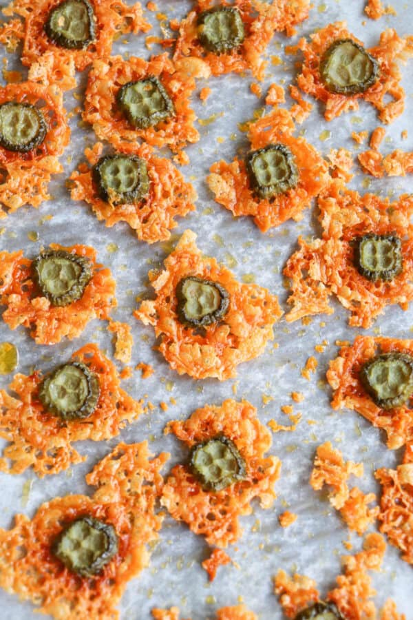 Close-up of baked cheese crisps with a slice of pickled green cucumber on each, resting on a parchment-lined baking sheet. The crisps are golden-brown and scattered randomly across the surface.
