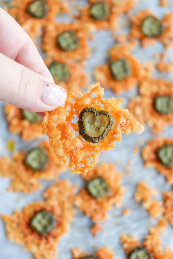 A hand holds a crispy cheese snack topped with a jalapeño slice. More similar snacks are scattered on a baking tray in the background.