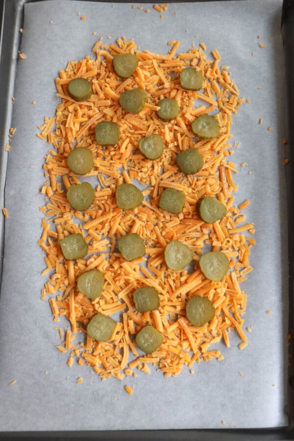 A baking sheet lined with parchment paper is covered with shredded cheddar cheese. Sliced pickles are evenly distributed on top of the cheese, ready to be baked.