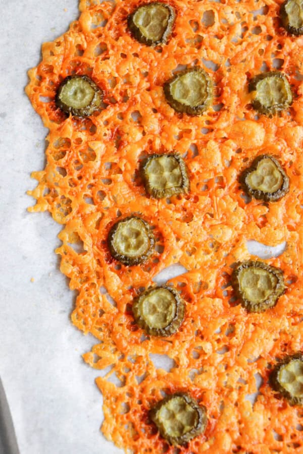 Close-up of crispy baked cheddar cheese slices with evenly spaced pickle chips on top, creating a lacy pattern. The cheese has a golden-brown color and the pickles are sliced thinly, sitting on parchment paper.