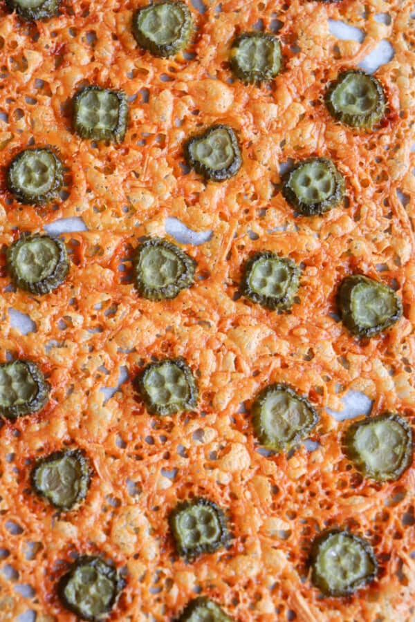 Close-up of baked cheese crisps topped with sliced pickles, featuring a crispy, golden-brown cheese texture arranged on a baking sheet. The pickles are evenly distributed, contrasting with the burnt orange hues of the melted cheese.
