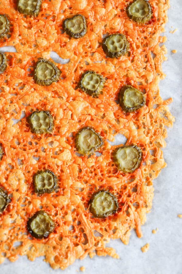 Close-up of crispy baked cheese slices topped with pickle slices on a sheet of parchment paper. The cheese is golden brown with lace-like edges, and the pickles are evenly distributed across the surface.