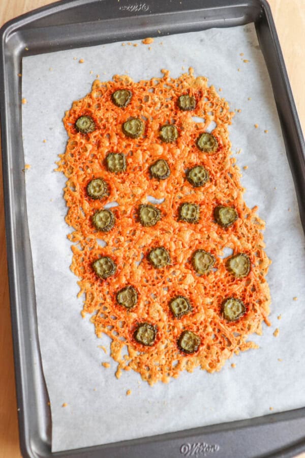 A baking sheet lined with parchment paper holds a flat layer of melted cheese crisps, topped with evenly spaced sliced pickles. The crispy cheese is golden brown with some toasty edges.