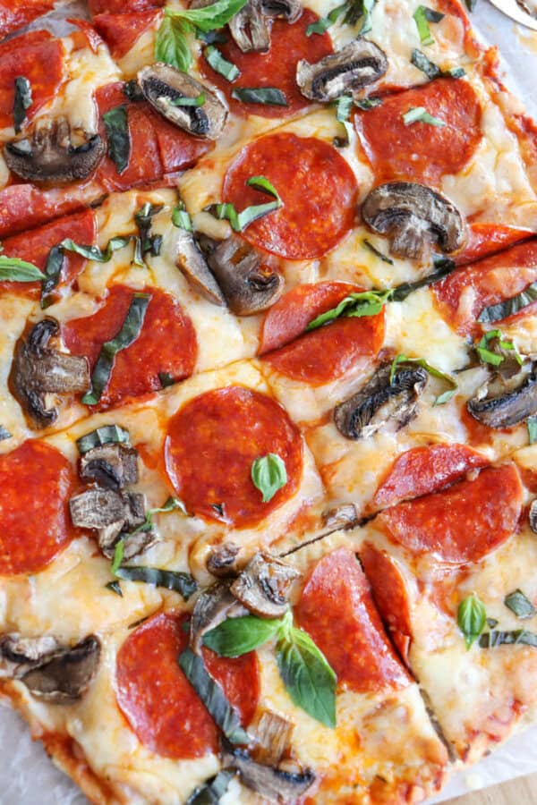 Close-up of a turkey crust pizza topped with pepperoni, mushrooms, and fresh basil leaves. The cheese is lightly browned, and the crust is golden. The pizza is sliced into squares, revealing a colorful and appetizing appearance.