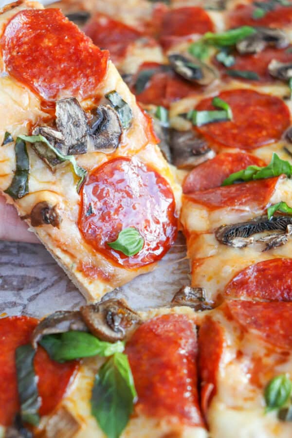 A hand holding a slice of turkey crust pizza topped with pepperoni, mushrooms, and fresh basil. The pizza is cut into squares on a wooden surface, with a golden-brown crust and melted cheese. Other slices are visible in the background.