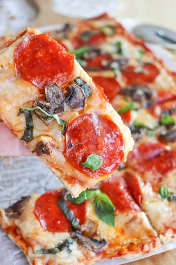 Close-up of a hand holding a slice of pizza topped with pepperoni, mushrooms, and fresh basil. The pizza appears to have a cheesy crust with a golden brown color. More pizza slices are visible in the background.