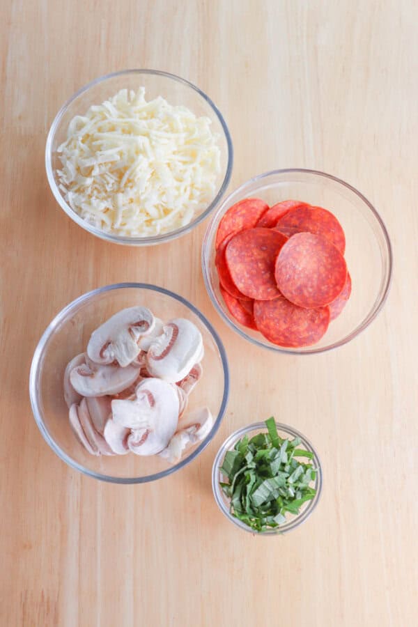 Four glass bowls on a wooden surface contain shredded cheese, pepperoni slices, sliced mushrooms, and chopped green herbs.