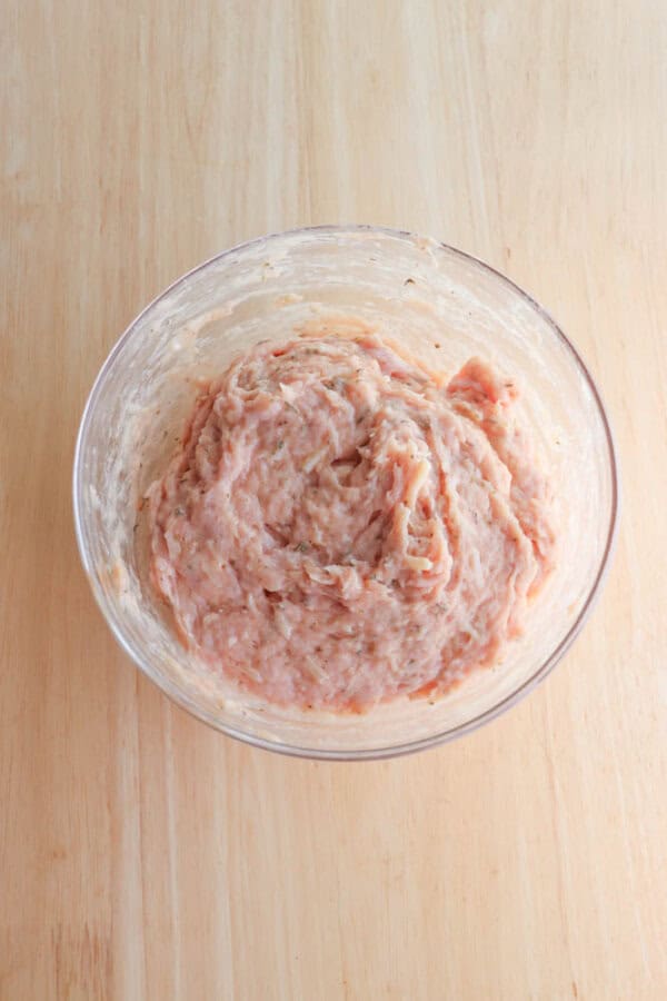 A clear glass bowl filled with raw, mixed ground meat rests on a light wooden surface. The meat appears finely blended, with a pinkish hue and a slightly sticky texture.