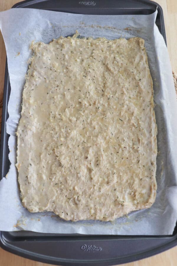A baking sheet lined with parchment paper holds a thin, raw dough spread evenly across its surface. The dough appears seasoned with herbs and is ready for baking. The baking sheet rests on a light wooden table.