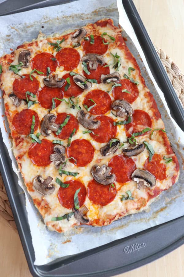 Rectangular homemade pizza topped with pepperoni, sliced mushrooms, and fresh basil, on a parchment-lined baking tray. The crust is golden brown and the cheese is melted and bubbly. The tray rests on a woven mat.