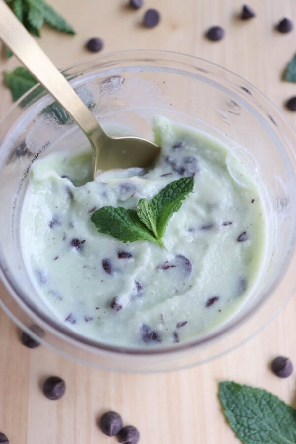 A bowl of mint chocolate chip ice cream with a gold spoon. The ice cream is light green with dark chocolate chips and topped with a fresh mint leaf. Scattered chocolate chips and mint leaves are around the bowl on a wooden surface.