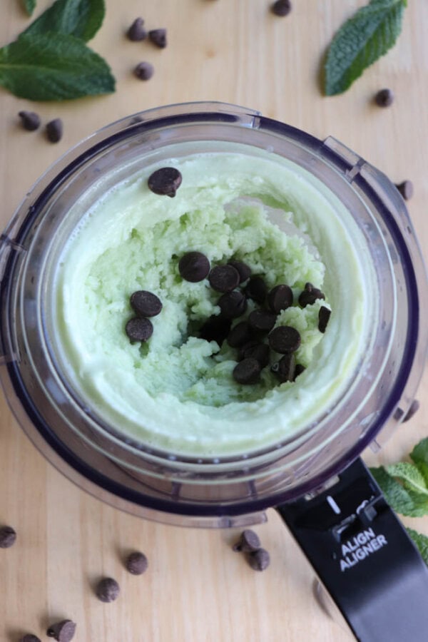 A top-down view of mint chocolate chip ice cream in a ninja creami cup. The ice cream is light green and topped with dark chocolate chips. Mint leaves and scattered chocolate chips surround the blender on a wooden surface.