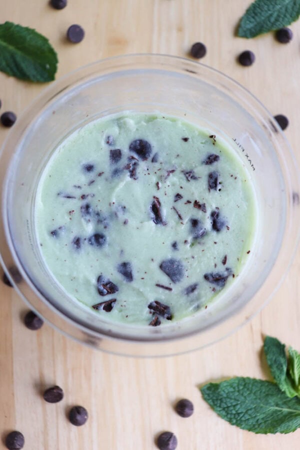 A ninja pint filled with mint chocolate chip ice cream, featuring a light green base and dark chocolate chunks. Surrounding the bowl are scattered chocolate chips and a few fresh mint leaves on a wooden surface.