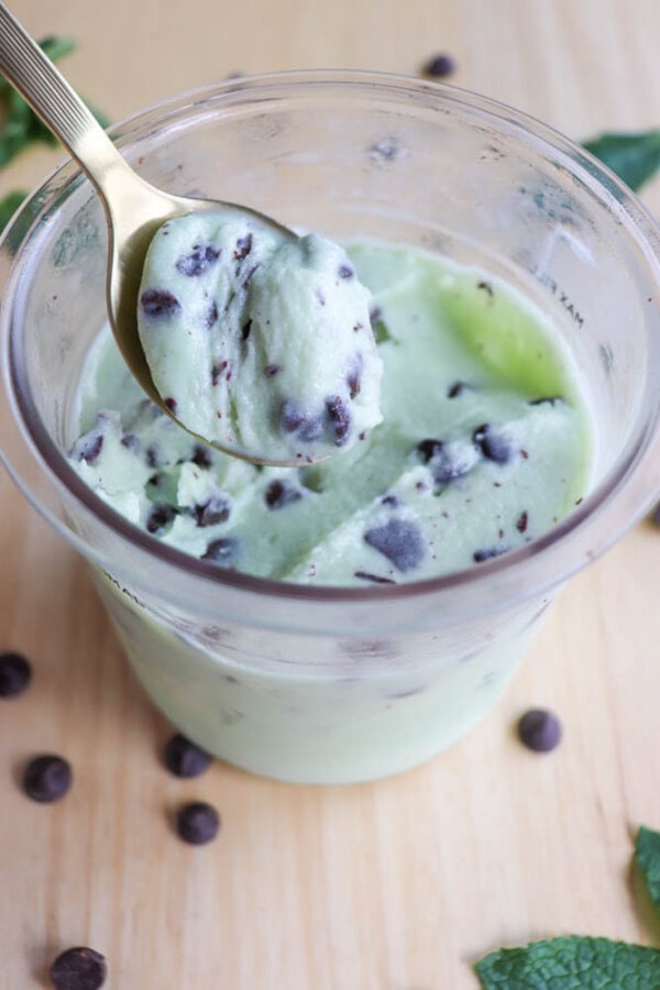 A glass jar filled with mint chocolate chip ice cream sits on a wooden surface. A spoon with a scoop of the ice cream rests above the jar, showcasing its creamy texture and chocolate chips. Mint leaves and chocolate chips are scattered around.