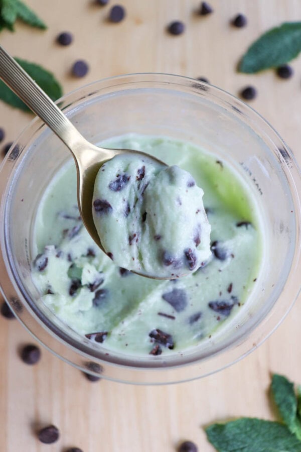 A spoonful of mint chocolate chip ice cream is held above a container filled with the same ice cream. Chocolate chips are scattered on the wooden surface nearby, and mint leaves are visible at the top corners.