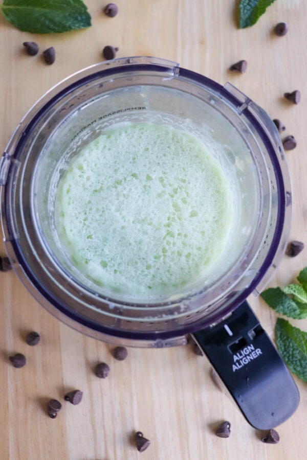A top view of a ninja cup filled with light green, frothy liquid, surrounded by scattered chocolate chips and fresh mint leaves on a wooden surface.