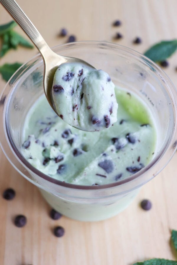 A spoon holds a scoop of mint chocolate chip ice cream above a small cup filled with more of the same ice cream. Chocolate chips are visible throughout, and a few chips are scattered on the wooden surface around the cup.