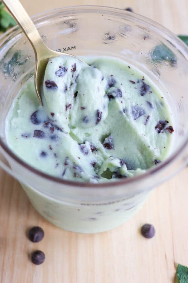 A clear plastic pint holds creamy mint chocolate chip ice cream. A gold spoon is scooping the ice cream, revealing visible dark chocolate chips. A few chocolate chips are scattered on the wooden surface around the container.