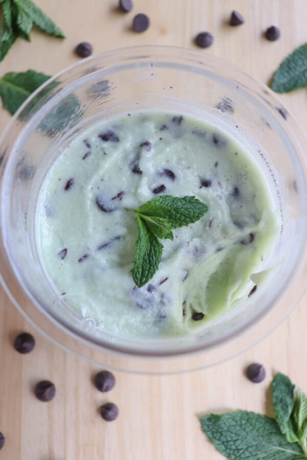 A bowl of mint chocolate chip ice cream, garnished with a fresh mint leaf. Chocolate chips are scattered around the ice cream and on the wooden surface. Mint leaves are also spread around for decoration.