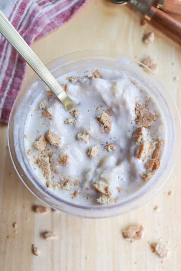 A bowl of creamy dessert topped with crumbled cookies. A spoon rests in the bowl. The background features a light-colored wooden surface and part of a red and white striped cloth.