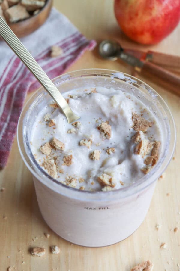 A clear cup filled with creamy yogurt topped with crumbled pieces of biscuit. A spoon is placed inside. A red apple, striped cloth, and an ice cream scoop are blurred in the background on a wooden surface.