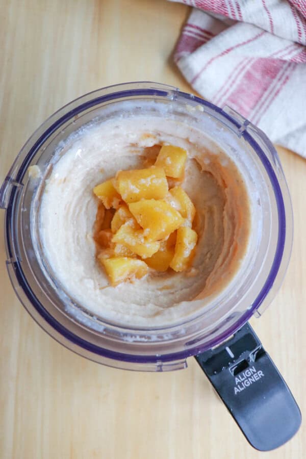 A food processor filled with creamy beige mixture with chunks of pineapple in the center. The appliance sits on a wooden surface, next to a folded red and white checkered cloth.