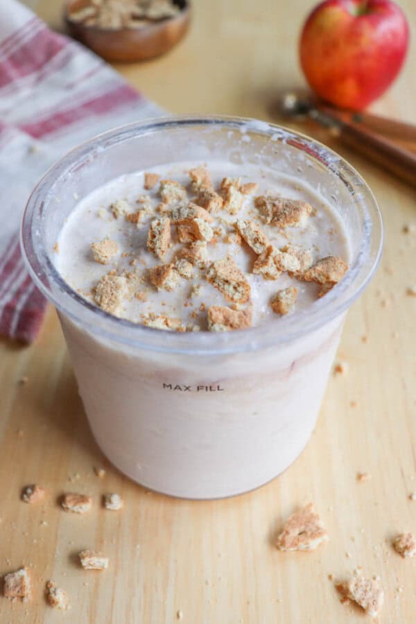A cup filled with creamy apple crumble ice cream topped with crumbled cookies sits on a wooden table. Nearby are an apple, a striped cloth, cinnamon sticks, and a small bowl with more cookie pieces.
