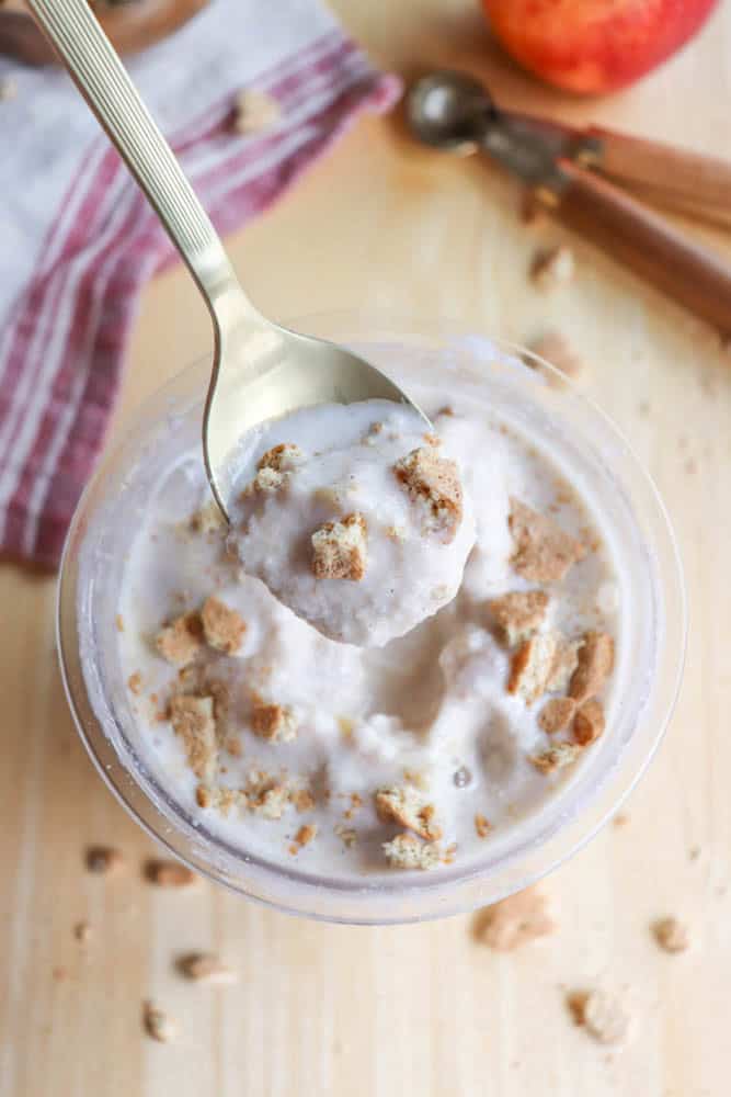 A spoon scooping a creamy dessert topped with crumbly pieces from a bowl. The light-colored dessert appears smooth with chunks mixed in. A red and white striped cloth, measuring spoons, and an apple partially visible in the background.