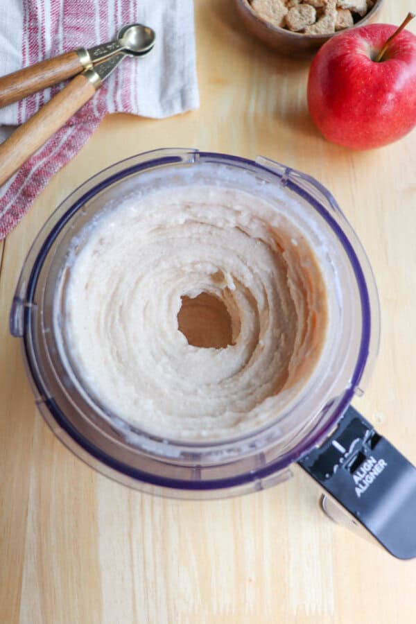 A food processor containing blended oats mixture on a wooden table. Nearby are a red apple, cereal pieces in a wooden bowl, and wooden measuring spoons on a red-striped cloth.