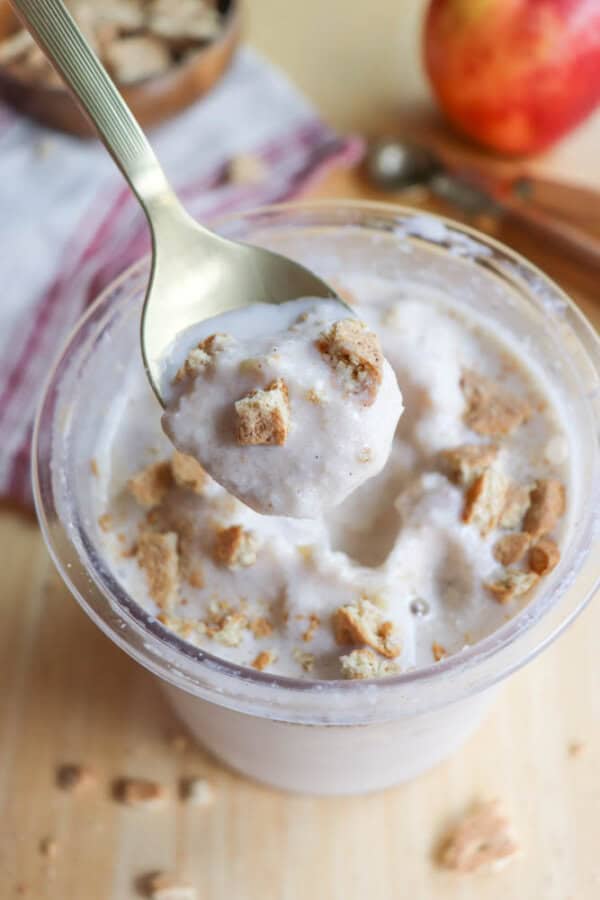 A spoon hovering over a cup of creamy dessert topped with crumbled cookies. A blurred apple and dish are in the background on a wooden surface.