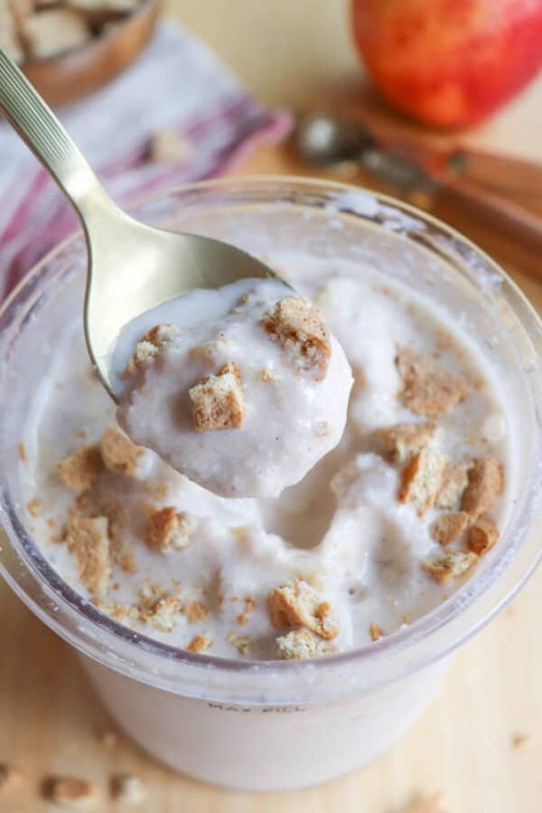 A spoonful of creamy dessert with crushed cookies is lifted from a clear cup. The dessert has a light, fluffy texture and is topped with cookie pieces. In the background, theres an apple and striped cloth on a wooden surface.