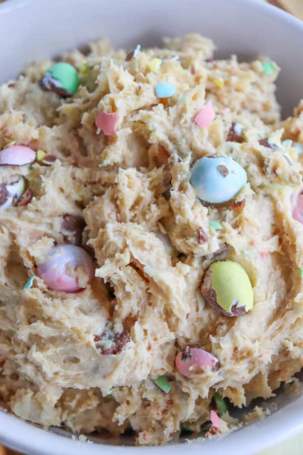 A bowl of cookie dough mixed with multicolored chocolate candies. The dough has a creamy texture with chunks of chocolate and pastel-colored candy pieces scattered throughout.