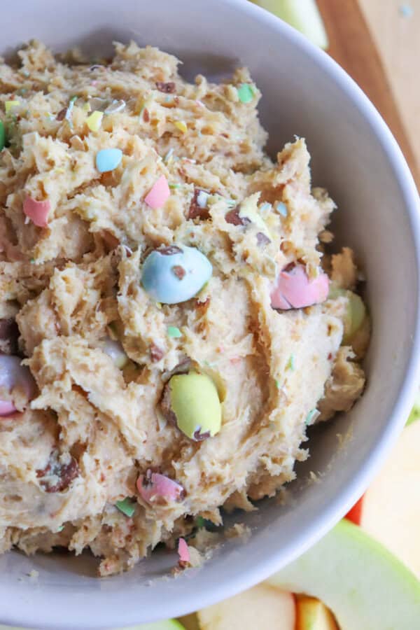 A bowl of cookie dough mixed with colorful candy-coated chocolate pieces. The dough is light brown, with visible candies in pastel colors like pink, blue, yellow, and green, offering a festive appearance.
