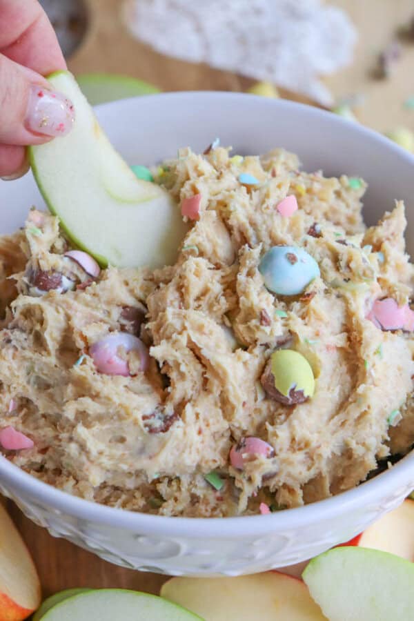 A person dips a slice of green apple into a bowl of cookie dough dip. The dip contains colorful candy-coated chocolates and is set on a wooden table. Apple slices are scattered around the bowl.