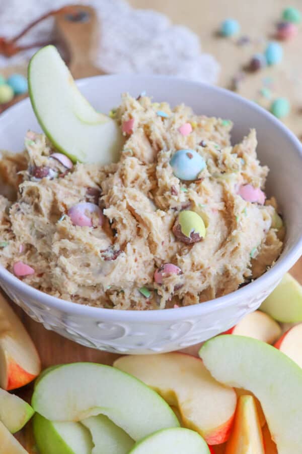 A bowl of light brown cookie dough dip with colorful candy pieces, garnished with an apple slice. Slices of red and green apples surround the bowl on a wooden surface.