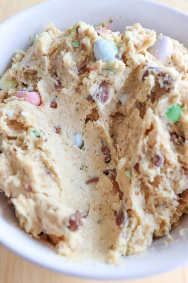 A bowl of cookie dough with colorful candy pieces mixed in. The dough is light beige and has a creamy, textured appearance. The candies are pastel-colored, adding a pop of color throughout the mixture. The bowl is placed on a light-colored surface.