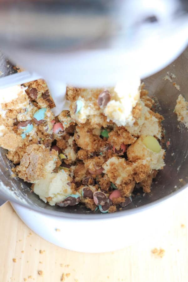 A mixing bowl with cookie dough containing chocolate chips and colorful candy pieces sits under a mixer. The dough is partially mixed, with chunks of ingredients visible. The mixer is placed on a light wooden surface.