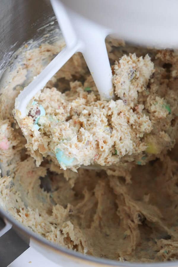 Cookie dough with colorful candy pieces is being mixed in a metal stand mixer bowl, with a white paddle attachment slightly lifted to show the doughs texture.