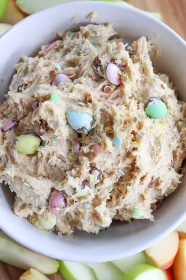 A bowl filled with cookie dough featuring colorful chocolate candy pieces. The dough has a creamy texture with visible candy pieces in pastel shades like pink, green, and yellow. Slices of apple are partially visible around the bowls edge.