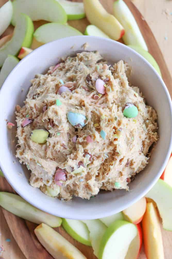A white bowl filled with creamy candy cookie dough, studded with colorful pastel candy pieces. The bowl is placed on a platter surrounded by slices of green and red apples on a wooden surface.