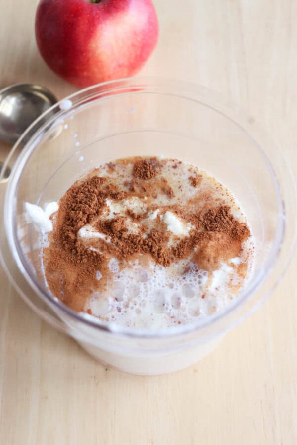 A clear plastic cup filled with a creamy mixture and topped with a layer of cinnamon. In the background, a red apple and a measuring spoon are visible on a light wooden surface.