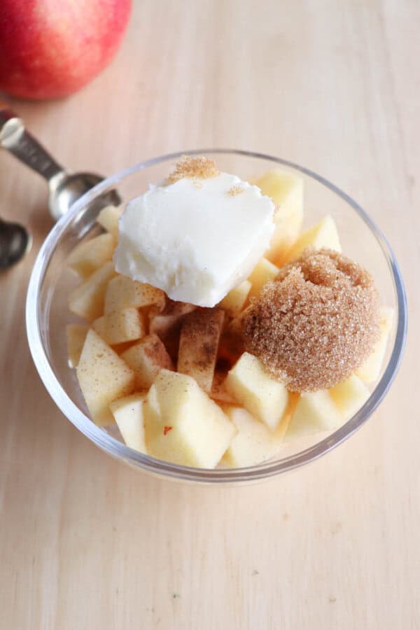 A clear bowl filled with diced apples topped with a scoop of butter, a mound of brown sugar, and a sprinkle of cinnamon. Spoons are in the background on a wooden surface, with a red apple partially visible.