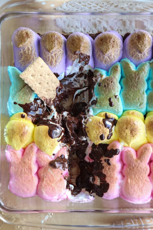 A baking dish filled with colorful marshmallow Peeps lined up in rows. Melted chocolate and a piece of graham cracker are in the center, resembling a smores dip.