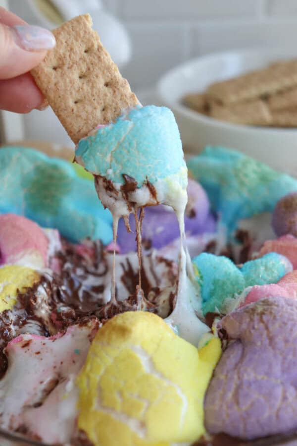 A hand dipping a graham cracker into melted chocolate and colorful marshmallow fluff. The marshmallow is swirled in pastel shades of pink, yellow, blue, and purple, creating a vibrant, gooey, and tempting treat.