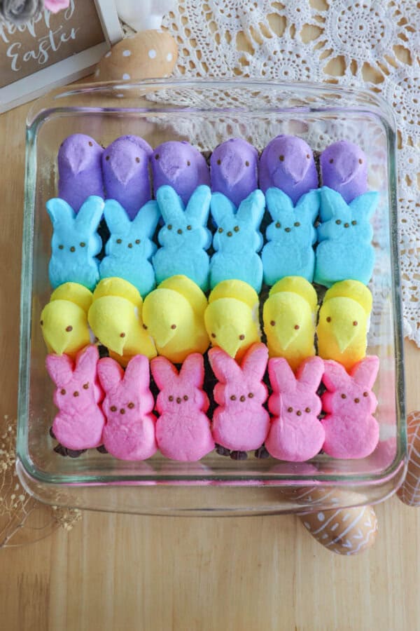 A glass dish filled with colorful marshmallow treats shaped like chicks and bunnies. The chicks are purple, blue, and yellow, while the bunnies are pink. The dish is on a wooden surface with decorative items nearby.