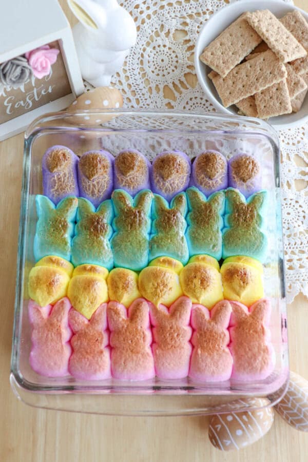 A dish of colorful marshmallow bunnies arranged in rows, lightly toasted on top. Next to it are graham crackers on a doily and decorated Easter eggs. A framed Easter decoration is in the background.